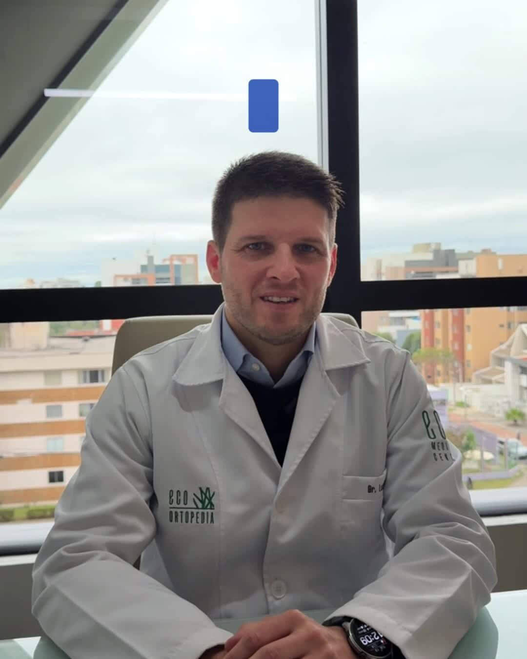 Dr. Luiz Müller Avila de jaleco, sentado diante de uma janela, gravando um vídeo