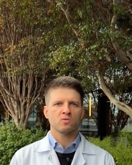 Dr. Luiz Müller Avila de jaleco, em pé num jardim, gravando um vídeo